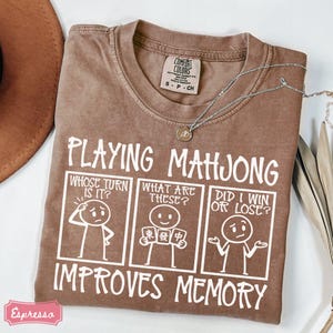 May include: A brown t-shirt featuring white text that reads Playing Mahjong Improves Memory. Below the text are three comic panels showing a stick figure asking Whose Turn Is It, What Are These, and Did I Win Or Lose. The shirt has a Comfort Colors tag.
