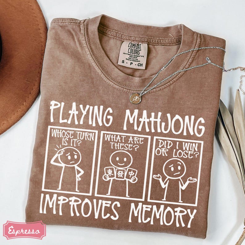 May include: A brown t-shirt featuring white text that reads Playing Mahjong Improves Memory. Below the text are three comic panels showing a stick figure asking Whose Turn Is It, What Are These, and Did I Win Or Lose. The shirt has a Comfort Colors tag.