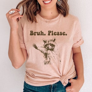 May include: A light pink t-shirt featuring a brown raccoon graphic and the text "Bruh. Please."