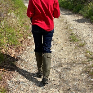 May include: A person wearing a red fleece jacket, dark blue jeans, and olive green gaiters walks along a gravel path. The path is surrounded by green grass and trees, with sunlight filtering through the leaves.