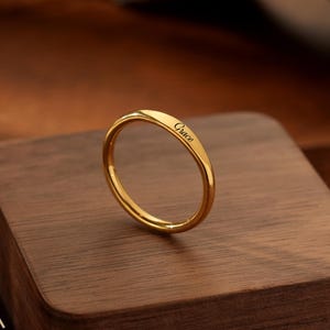 Può includere: Un sottile anello a fascia in metallo dorato lucido con il nome Grace inciso in corsivo sulla superficie esterna. L'anello presenta un profilo liscio e arrotondato con una finitura metallica riflettente dal design semplice ed essenziale.