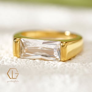 May include: A gold ring with a clear rectangular stone set in the center. The ring is thick and has a polished finish.
