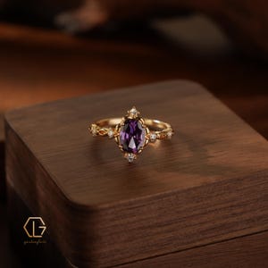 May include: A gold ring featuring a central oval-cut purple gemstone surrounded by small clear stones. The band has a decorative metalwork design with additional small clear stones set along the sides. The logo text "GL" is visible in the corner.