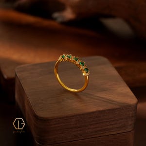 May include: A gold ring featuring a row of alternating small round green gemstones and clear white stones set along the top half of the band. The ring has a polished metallic finish and a thin, circular band design that sits upright on a surface.