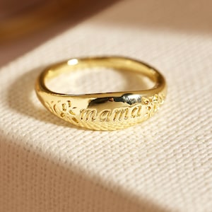 May include: A gold ring featuring the word "mama" engraved on the front. The ring has a floral pattern on the sides. The ring is displayed on a neutral surface. A piece of jewellery.
