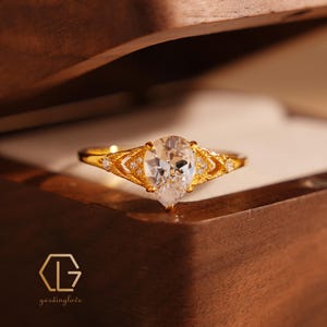 Può includere: Un anello d'oro con una pietra preziosa trasparente a forma di pera e dettagli intricati. L'anello è posto in una scatola di legno con il testo "gardinglove" sul fondo.