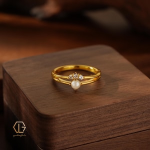 Puede incluir: Un anillo de tono dorado con un diseño de banda dividida. El centro del anillo está adornado con una perla blanca única colocada debajo de un pequeño engaste arqueado que contiene tres piedras claras y brillantes en fila.