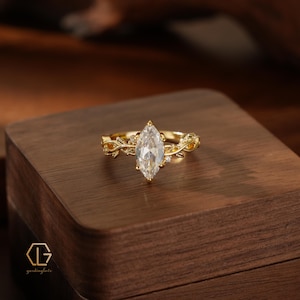 May include: A gold ring featuring a central marquise-cut clear gemstone. The band is designed with a textured vine and leaf motif, accented by small, round, clear stones embedded within the metal foliage. The ring is displayed on a wooden surface.