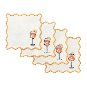 Puede incluir: Juego de cuatro servilletas de cóctel de lino blanco con bordes bordados de color naranja. Cada servilleta presenta una imagen bordada de un vaso de zumo de naranja con una rodaja de naranja en el borde.