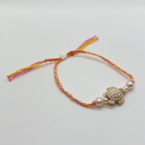 Puede incluir: Pulsera hecha a mano con un pequeño dije de tortuga blanquecina y dos perlas de imitación. El cordón de la pulsera es una mezcla trenzada de hilos rosas, amarillos y blancos, creando una estética colorida y playera.
