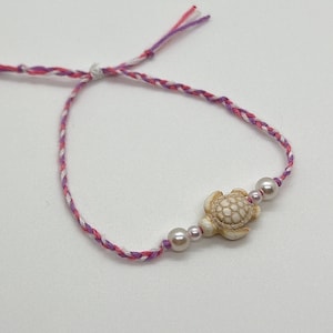 Puede incluir: Un brazalete de cuerda trenzada rosa, blanco y rojo con un dije de tortuga blanca y dos cuentas de perla blanca.