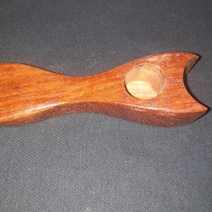 May include: A wooden pipe with a rounded bowl and a small, round hole in the center. The pipe is made of a reddish-brown wood and has a smooth finish.