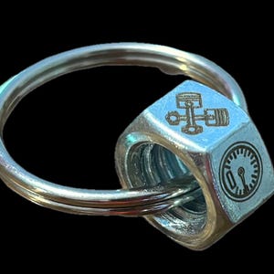 May include: A silver metal keychain with a hexagonal nut attached to a ring. The nut has an engraved image of a car engine and a speedometer.
