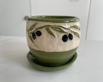 May include: A ceramic plant pot with a matching saucer, featuring a two-tone design with a solid green base and a textured cream-colored upper section decorated with hand-painted green olive branches and dark blue olives.