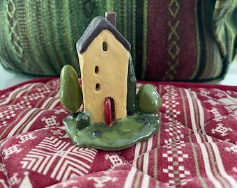 May include: A small ceramic house figurine with a pale yellow facade, a dark brown roof, and a red door. The structure is set on a green base with two green trees on either side. The house features three small vertical windows on the front.