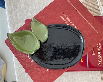 May include: A ceramic dish featuring a large, glossy black oval base with two attached, smaller, light green leaf-shaped bowls. The surface of the black section has a textured, slightly uneven finish, while the green leaves have a smooth, reflective glaze.