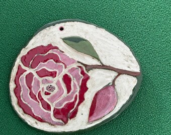 May include: A circular ceramic ornament featuring a hand-painted pink and deep red rose with green leaves on a white background. The piece has a glossy finish and a small hole at the top for hanging, with a dark grey edge border.