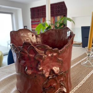 Op de afbeelding: Een cilindrische keramische vaas met een glanzend, dieprood glazuur. De buitenkant is voorzien van driedimensionale, sculpturale bladvormige applicaties die verticaal zijn gerangschikt. De rand van de vaas heeft een onregelmatige, golvende vorm.