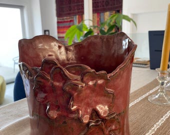 May include: A cylindrical ceramic vase with a glossy, deep red glaze. The exterior features three-dimensional, sculpted leaf-like appliques arranged vertically. The rim of the vase has an irregular, wavy edge, and the base is circular.