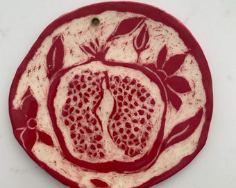 May include: A round, hand-formed ceramic wall plaque featuring a deep red glaze with a contrasting white, incised design of a halved pomegranate surrounded by leaves and flowers. A small circular hole is positioned at the top for hanging.