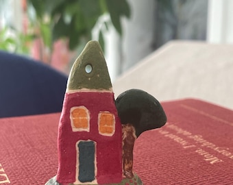 May include: A small, hand-painted ceramic house ornament with a pointed green roof, a magenta facade, two orange windows, and a blue door. A dark green tree with a brown trunk is attached to the side, and a small hole is at the top of the roof.