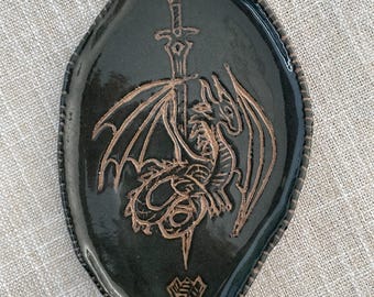 May include: A leaf-shaped ceramic trinket dish with a glossy black glaze. The surface features an etched design of a dragon coiled around a sword, with a small geometric symbol at the bottom. The edges are textured with a series of small indentations.