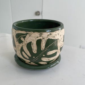 May include: A dark green ceramic planter with a matching saucer, featuring a carved, cream-colored monstera leaf pattern wrapping around the exterior. The pot has a smooth, glossy finish and a wide, circular opening with a thick, rounded rim.