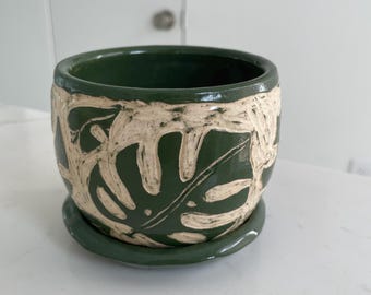 May include: A dark green ceramic planter with a matching saucer, featuring a carved, cream-colored monstera leaf pattern wrapping around the exterior. The pot has a smooth, glossy finish and a wide, circular opening with a thick, rounded rim.