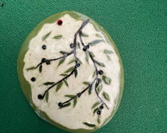 May include: An oval ceramic pendant with a glossy finish, featuring a cream-colored center painted with a dark brown branch, green leaves, and small black berries. The outer border is a solid olive green, and a small hole is visible at the top.