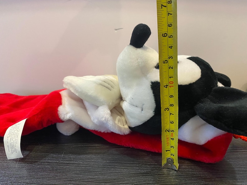 May include: A plush toy shaped like a cartoon character, featuring a black and white head, white gloves, and a red stocking. The toy measures approximately 6 inches in height.