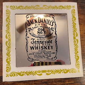 Könnte beinhalten: Rechteckiger Spiegel mit dem Logo der Marke Jack Daniel's Old No. 7. Er hat einen weißen Rahmen mit einem goldenen floralen Design. Der Spiegel zeigt den Text "Tennessee Sour Mash Whiskey".