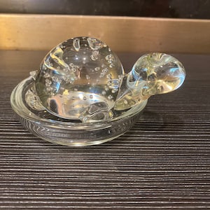 Puede incluir: Una figura de tortuga de cristal transparente con un caparazón lleno de burbujas, colocada en un cuenco de cristal a juego. La cabeza y las aletas de la tortuga también son de cristal transparente. Un artículo decorativo.