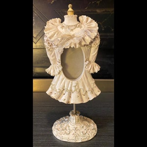 Pode incluir: Moldura decorativa em forma de vestido, em branco sujo. A moldura apresenta detalhes intrincados, incluindo acabamentos em renda, mangas com babados e detalhes dourados. Assenta numa base ornamentada, adicionando um toque vintage.