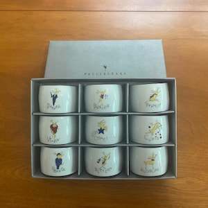 Pode incluir: Um conjunto de nove argolas de guardanapo de cerâmica branca, cada uma com um design de personagem de rena diferente e um nome. Os nomes incluem Dasher, Dancer, Prancer, Vixen, Comet, Cupid, Donner, Blitzen e Rudolph. As argolas de guardanapo estão em uma caixa cinza.