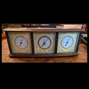 May include: A vintage wooden weather station with three analog gauges. The gauges display temperature, barometric pressure, and humidity. The faces are cream-colored with black numerals and hands, and the frame is dark brown.
