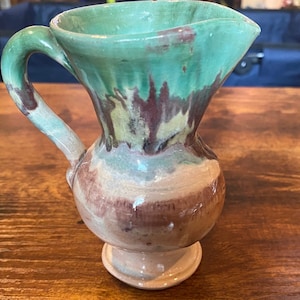 May include: A handcrafted ceramic pitcher with a green, brown, and beige glaze. The pitcher has a curved handle and a flared spout. The base is a small, round pedestal. The pitcher is likely used for serving beverages.