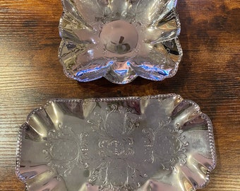 Puede incluir: Dos fuentes plateadas. La fuente superior es cuadrada con un borde con volantes. La fuente inferior es rectangular con un borde con volantes y un diseño floral ornamentado. Ambas fuentes son reflectantes y están sobre una superficie de madera.
