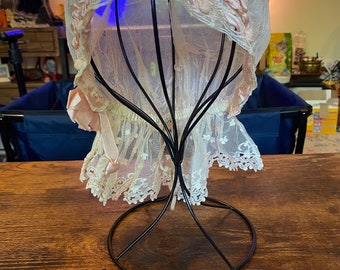May include: A vintage white lace bonnet with pink ribbon and floral accents, displayed on a black metal stand. The bonnet features a delicate net overlay and intricate lace detailing, showcasing a classic, elegant design.
