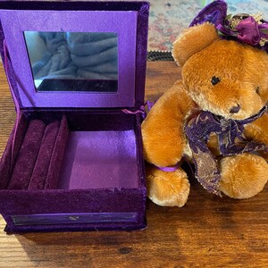 May include: A purple velvet jewellery box with a mirror inside and a brown teddy bear wearing a purple hat and scarf. The jewellery box is open, revealing a velvet interior with ring slots.