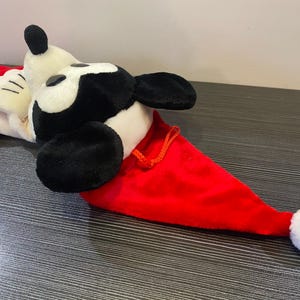 May include: A plush Mickey Mouse Christmas hat. The hat is red with a white pom-pom and a white and black Mickey Mouse head. The hat is lying on a dark surface.