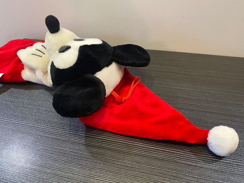 May include: A plush Mickey Mouse Christmas hat. The hat is red with a white pom-pom and a white and black Mickey Mouse head. The hat is lying on a dark surface.