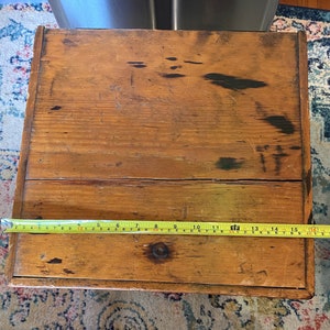 K&ouml;nnte beinhalten: Eine rechteckige Holzkiste mit nat&uuml;rlicher Oberfl&auml;che, die Gebrauchsspuren und Alter aufweist. Die Kiste wird mit einem gelben Ma&szlig;band gemessen, etwa 40 cm lang.