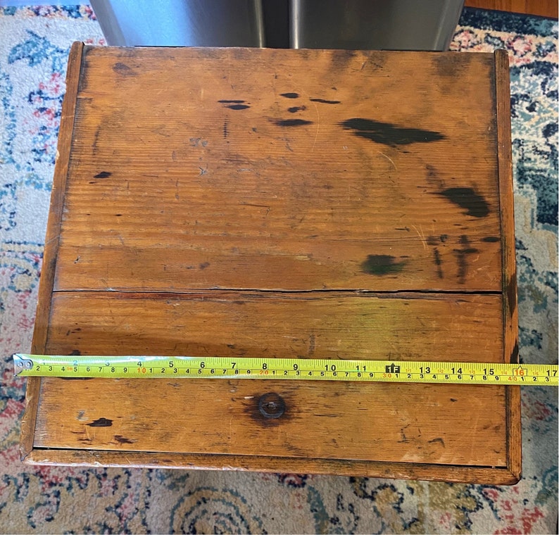 K&ouml;nnte beinhalten: Eine rechteckige Holzkiste mit nat&uuml;rlicher Oberfl&auml;che, die Gebrauchsspuren und Alter aufweist. Die Kiste wird mit einem gelben Ma&szlig;band gemessen, etwa 40 cm lang.