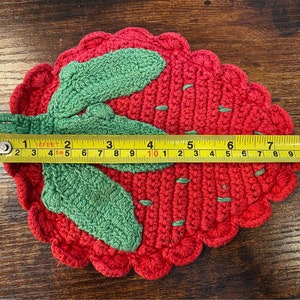 K&ouml;nnte beinhalten: Handgefertigter Erdbeer-Untersetzer aus H&auml;kelarbeit. Der Untersetzer ist rot mit gr&uuml;nen Bl&auml;ttern und hat einen Durchmesser von ca. 18 cm. Der Untersetzer ist aus Garn gefertigt und hat einen gewellten Rand.