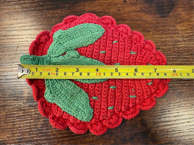 K&ouml;nnte beinhalten: Handgefertigter Erdbeer-Untersetzer aus H&auml;kelarbeit. Der Untersetzer ist rot mit gr&uuml;nen Bl&auml;ttern und hat einen Durchmesser von ca. 18 cm. Der Untersetzer ist aus Garn gefertigt und hat einen gewellten Rand.