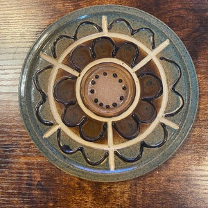 May include: A ceramic serving dish with a floral design. The dish features a central well surrounded by a series of petal-shaped compartments. The colors include shades of blue, brown, and cream, with dark brown accents. The dish is circular and appears handmade.
