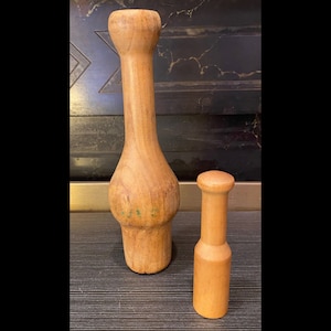 Peut inclure: Deux ustensiles de cuisine en bois. L'un est un pilon long, brun clair, avec une section m&eacute;diane renfl&eacute;e et un dessus arrondi. L'autre est un pilon cylindrique plus petit avec un dessus arrondi. Les deux sont en bois et ont une finition lisse.