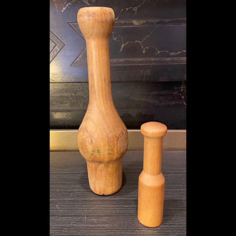Peut inclure: Deux ustensiles de cuisine en bois. L'un est un pilon long, brun clair, avec une section m&eacute;diane renfl&eacute;e et un dessus arrondi. L'autre est un pilon cylindrique plus petit avec un dessus arrondi. Les deux sont en bois et ont une finition lisse.