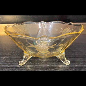 May include: A vintage, yellow-tinted glass bowl with a scalloped rim and three delicate feet. The bowl features etched floral designs, adding a touch of elegance. This decorative piece is ideal for serving or display.