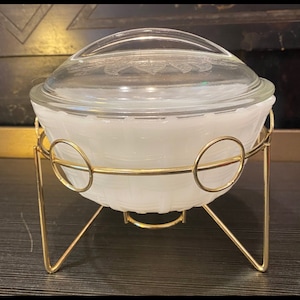 May include: A white, oval-shaped serving dish with a clear glass lid, set in a gold-colored metal stand. The dish has a basketweave pattern, and the lid features a decorative etched design. The stand has three legs.